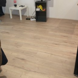 Grzegorz Karbowiak - Jasne panele podłogowe ułożone w salonie, widoczny fragment stolika z napojem, szafka RTV z telewizorem i routerem.