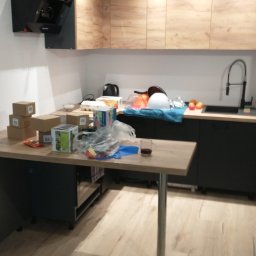 Grzegorz Karbowiak - Wnętrze kuchni w trakcie montażu, widoczne szafki z drewnopodobnym frontem, czarne szafki dolne, na blacie kartony, opakowania i narzędzia, co sugeruje trwające prace wykończeniowe.