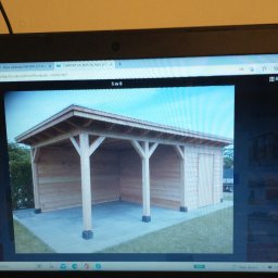 Grzegorz Karbowiak - Drewniana wiata garażowa z płaskim dachem i boczną ścianą z desek, widoczna na ekranie laptopa.