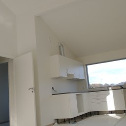 Grzegorz Karbowiak - Wnętrze w trakcie wykańczania: biała kuchnia z czarnym blatem, widoczne rury wentylacyjne, podłoga zabezpieczona folią ochronną z nadrukiem, drzwi otwarte do sąsiedniego pomieszczenia.