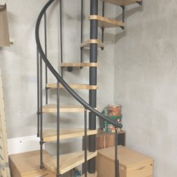 Grzegorz Karbowiak - Kompaktowe schody spiralne z drewnianymi stopniami i czarną metalową konstrukcją, umieszczone w pomieszczeniu z surowymi ścianami, obok biurek z szufladami i puszkami farb.