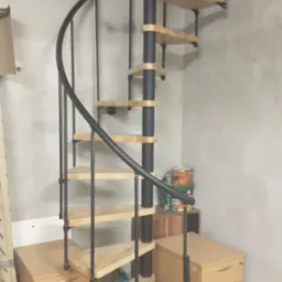 Kompaktowe schody spiralne z drewnianymi stopniami i czarną metalową konstrukcją, umieszczone w pomieszczeniu z surowymi ścianami, obok biurek z szufladami i puszkami farb.