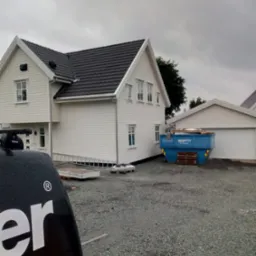 Nowo wybudowany, biały dom z czarnym dachem, widoczny z zewnątrz, z kontenerem na gruz obok garażu, elementy konstrukcyjne leżą na podłożu z grysu, widoczny fragment samochodu z bagażnikiem dachowym.