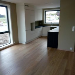 Grzegorz Karbowiak - Wnętrze nowo wykończonej kuchni z białymi szafkami i ciemnym blatem, widok na okno z widokiem na zewnątrz, drewniana podłoga.