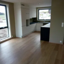 Wnętrze nowo wykończonej kuchni z białymi szafkami i ciemnym blatem, widok na okno z widokiem na zewnątrz, drewniana podłoga.