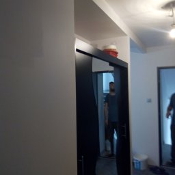 Grzegorz Karbowiak - Wnętrze mieszkania w trakcie remontu, widoczna ściana po malowaniu, drzwi przesuwne do garderoby z ciemną ramą, w tle mężczyzna i wiadro z farbą.