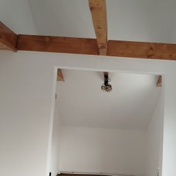 Grzegorz Karbowiak - Wnętrze po remoncie: białe ściany, drewniane belki stropowe, widoczny fragment podłogi i instalacji. Minimalistyczny design z surowym oświetleniem.