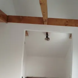 Wnętrze po remoncie: białe ściany, drewniane belki stropowe, widoczny fragment podłogi i instalacji. Minimalistyczny design z surowym oświetleniem.