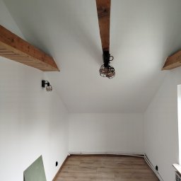 Grzegorz Karbowiak - Białe wnętrze poddasza z drewnianymi belkami stropowymi, lampami w stylu industrialnym i panelami podłogowymi. Na podłodze płyta karton gips.