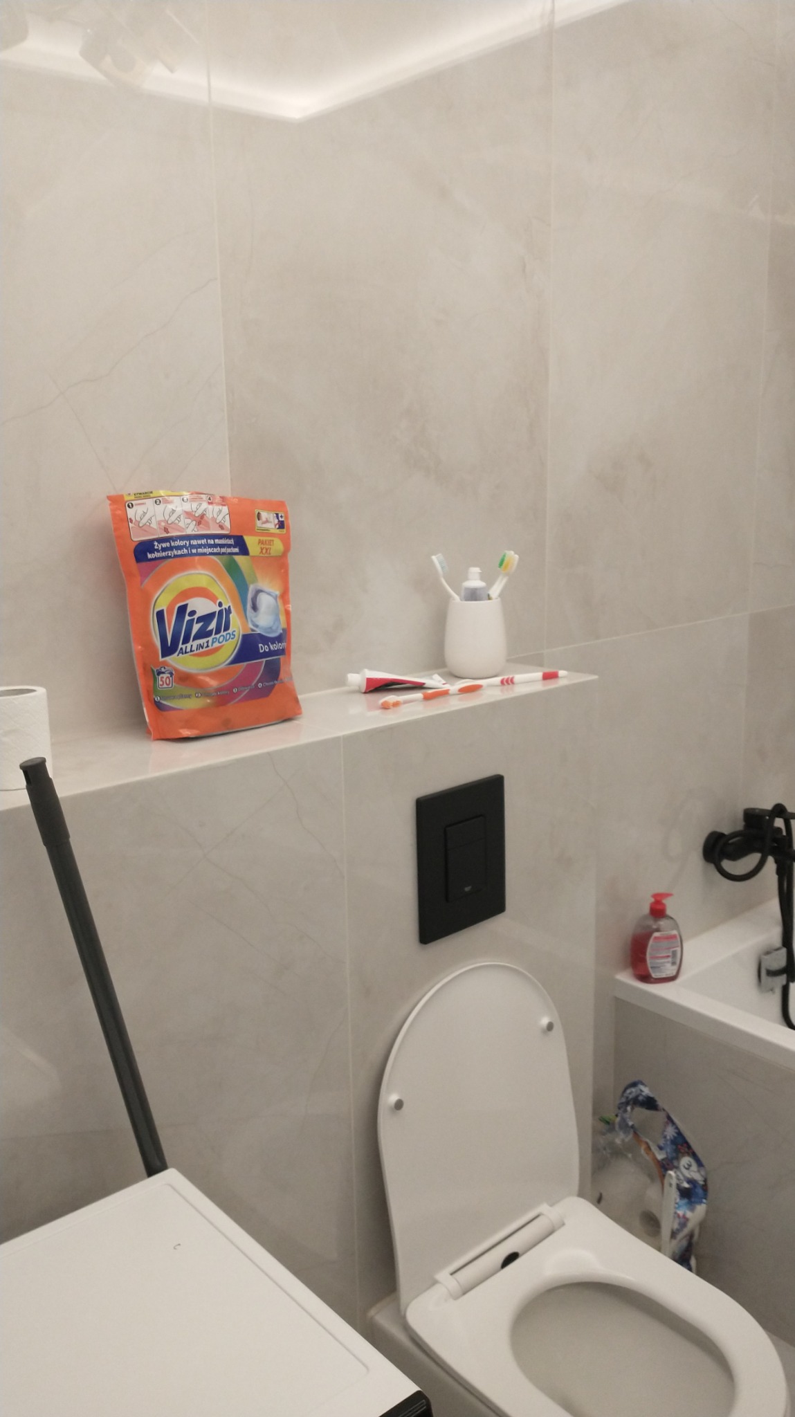 Jasna łazienka z toaletą, na półce leżą środki czystości i przybory toaletowe, widoczny fragment pralki.