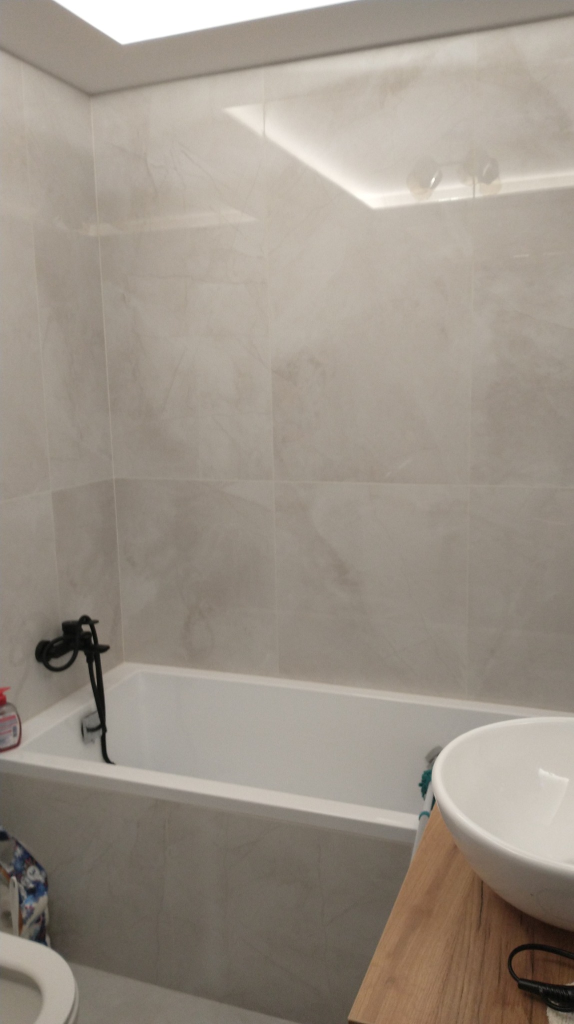 Wanna z czarną armaturą, umywalka nablatowa na drewnianym blacie w łazience wykończonej płytkami imitującymi marmur, z oświetleniem sufitowym.