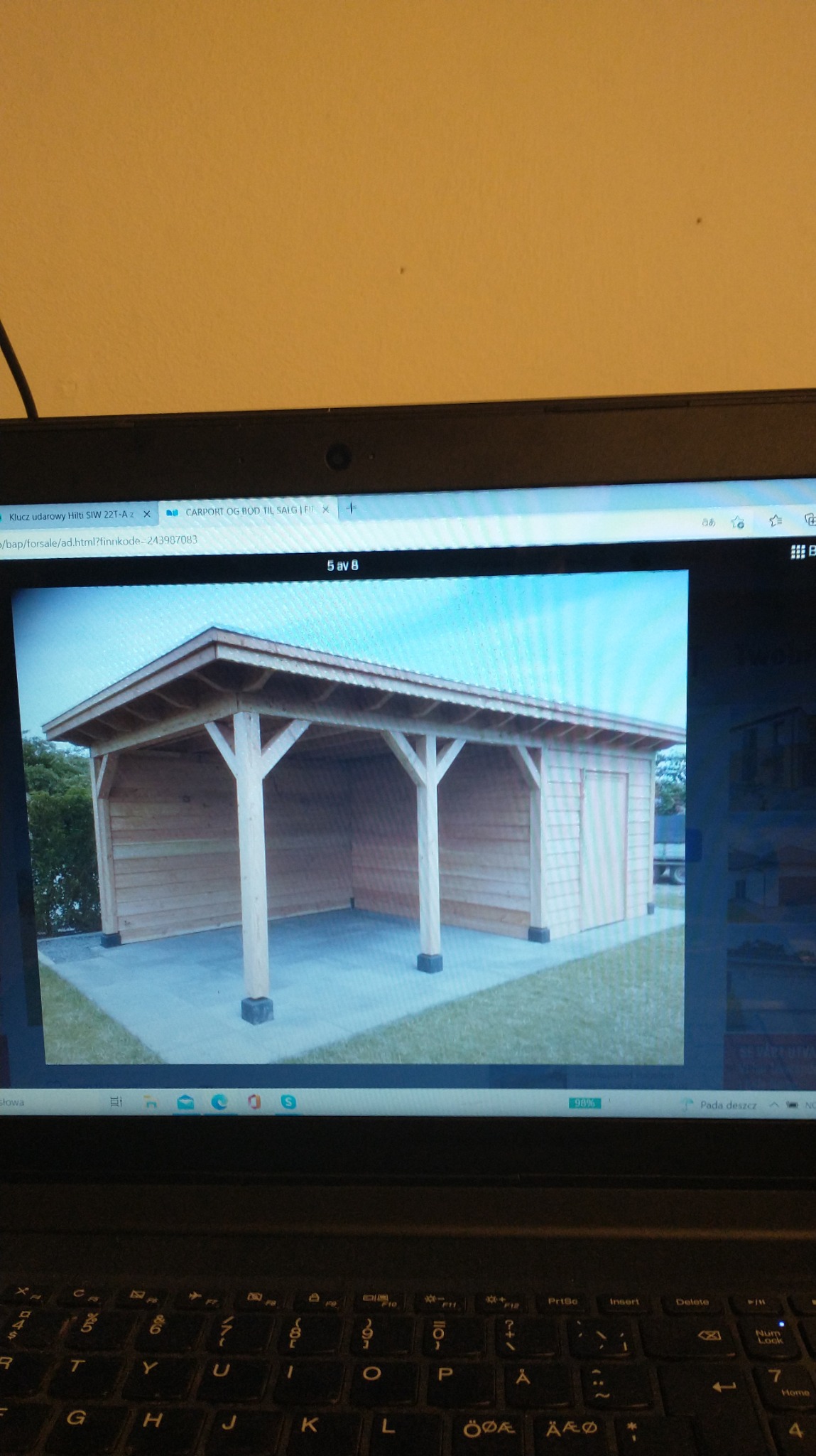 Drewniana wiata garażowa z płaskim dachem i boczną ścianą z desek, widoczna na ekranie laptopa.