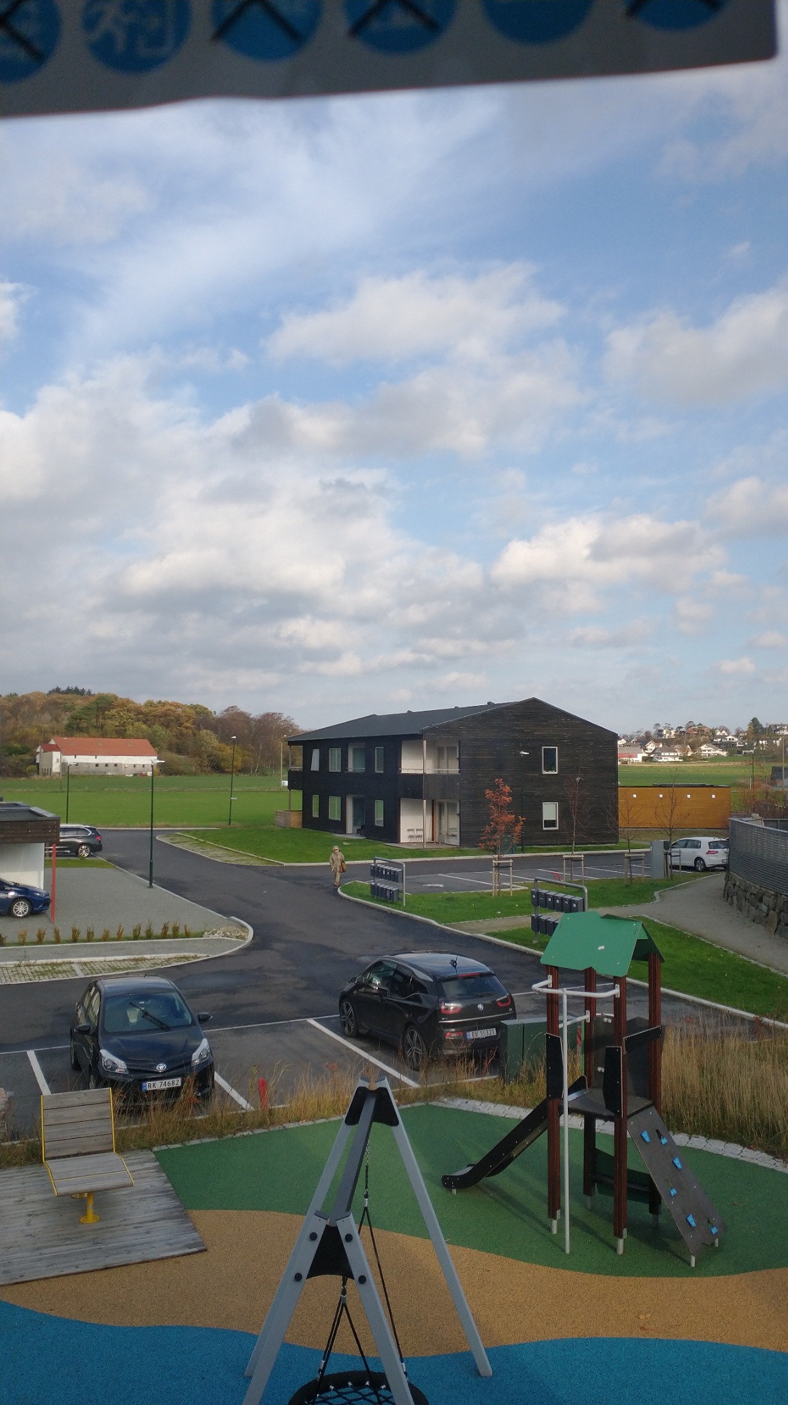 Dwupiętrowy budynek z ciemnego drewna z białymi elementami, widziany z placu zabaw z huśtawką typu bocianie gniazdo i zjeżdżalnią, zaparkowane samochody, zielony trawnik i pochmurne niebo w tle.