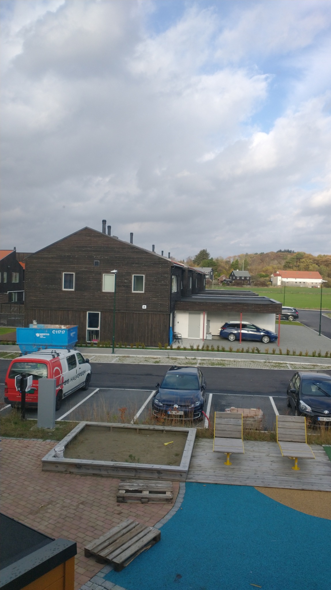 Dwupiętrowy budynek mieszkalny z ciemnego drewna z parkingiem, piaskownicą i ławkami na pierwszym planie, pochmurne niebo w tle.