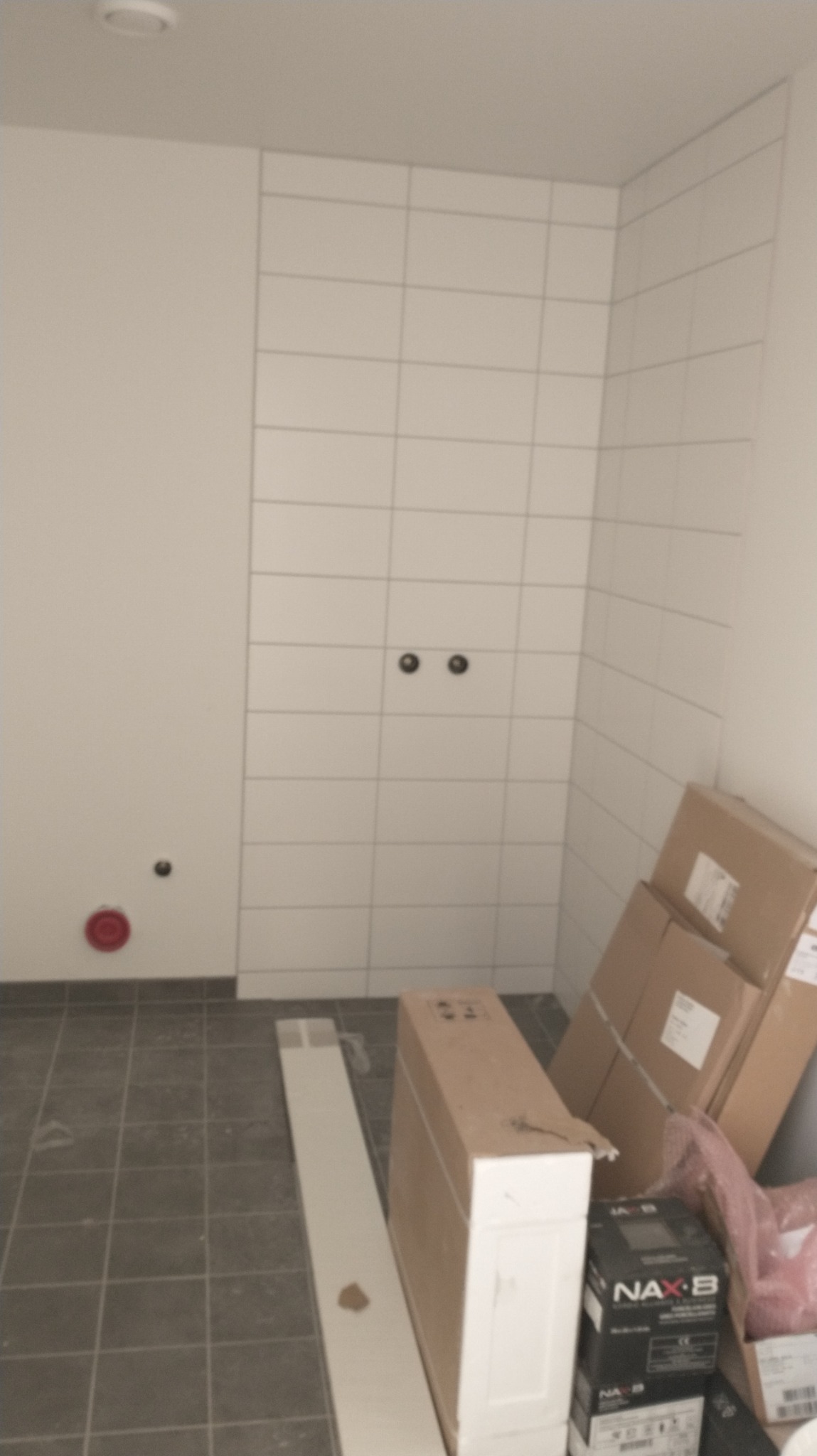 Ściana wyłożona białymi, prostokątnymi płytkami z widocznymi przyłączami wodno-kanalizacyjnymi; na podłodze szare płytki, kartony i biała listwa przypodłogowa.