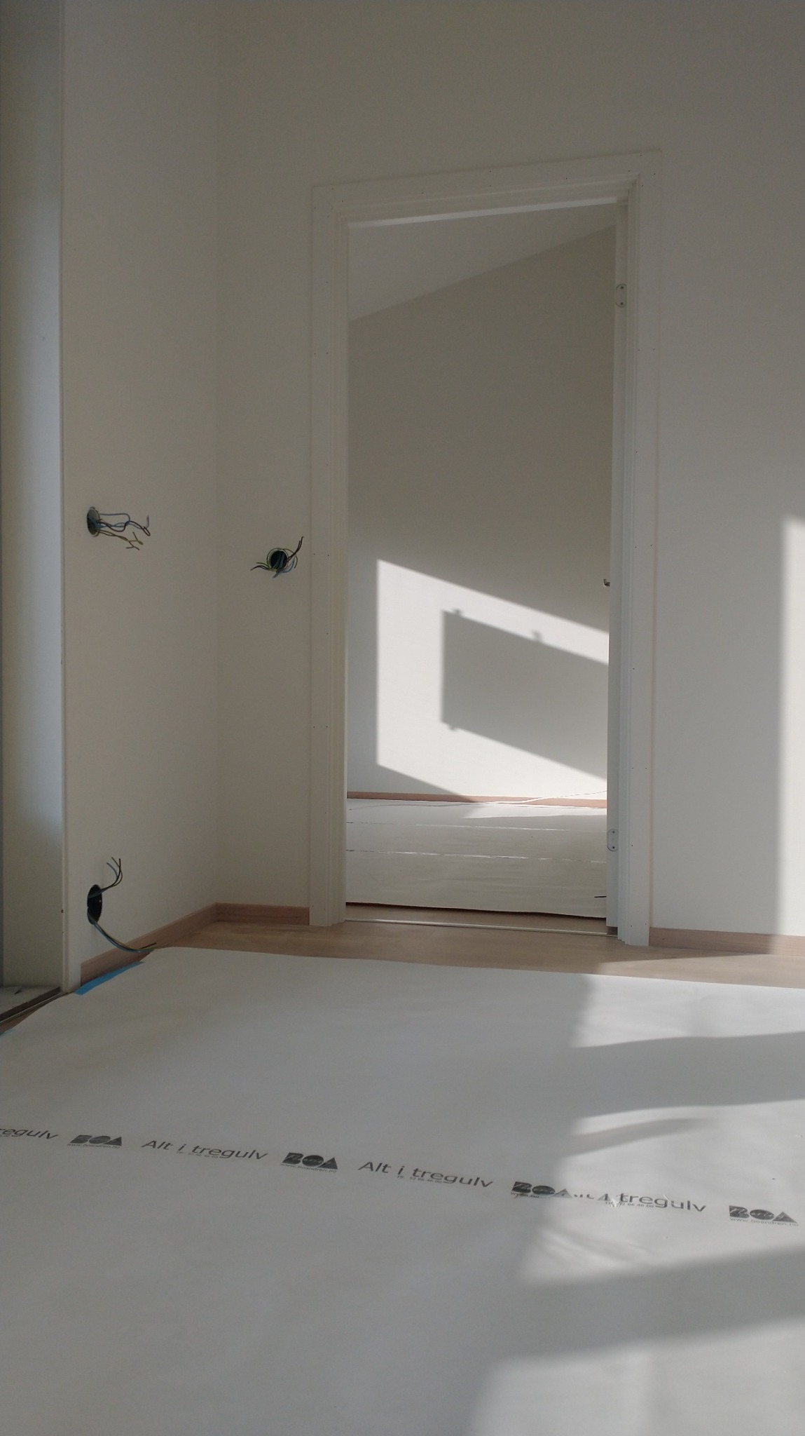 Wnętrze w trakcie prac wykończeniowych: widoczne przewody elektryczne wystające ze ściany, zabezpieczona podłoga folią ochronną z nadrukiem, oraz białe ściany i ościeżnica drzwiowa z widokiem...