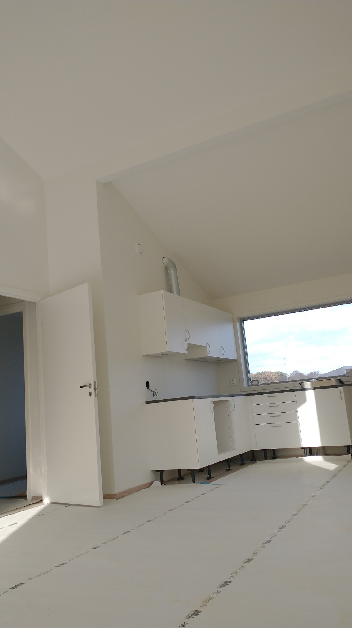 Wnętrze w trakcie wykańczania: biała kuchnia z czarnym blatem, widoczne rury wentylacyjne, podłoga zabezpieczona folią ochronną z nadrukiem, drzwi otwarte do sąsiedniego pomieszczenia.