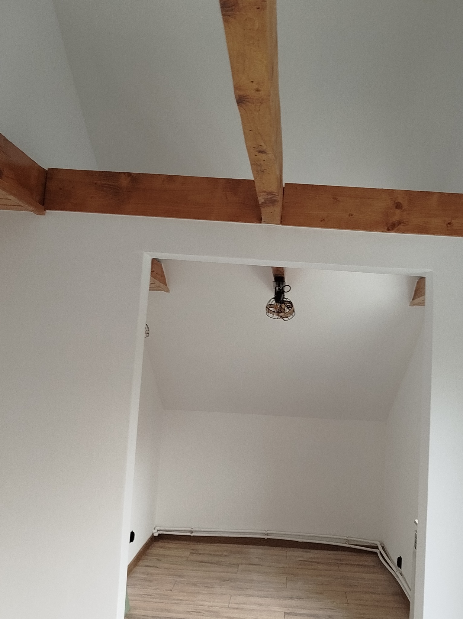 Wnętrze po remoncie: białe ściany, drewniane belki stropowe, widoczny fragment podłogi i instalacji. Minimalistyczny design z surowym oświetleniem.