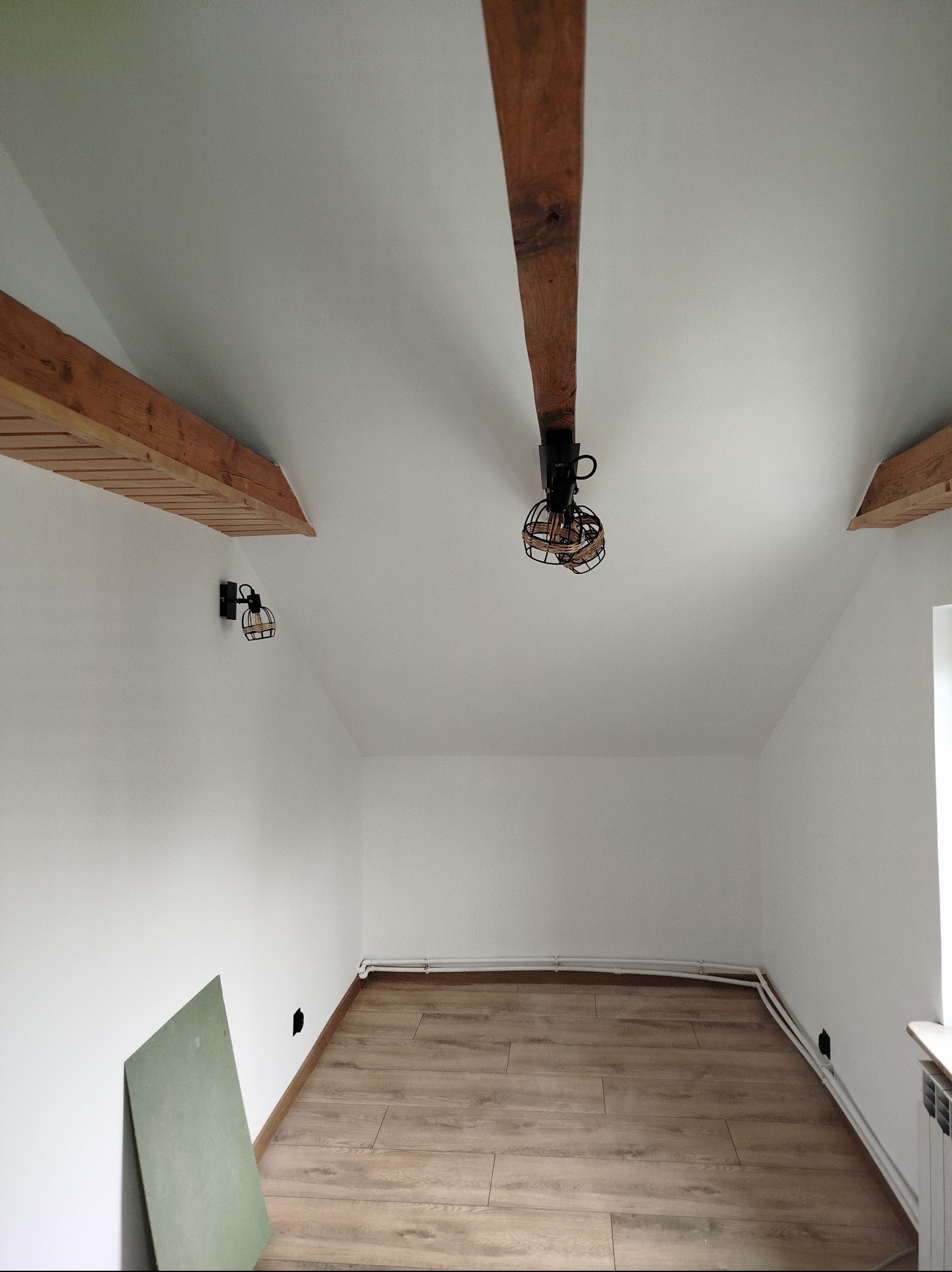Białe wnętrze poddasza z drewnianymi belkami stropowymi, lampami w stylu industrialnym i panelami podłogowymi. Na podłodze płyta karton gips.