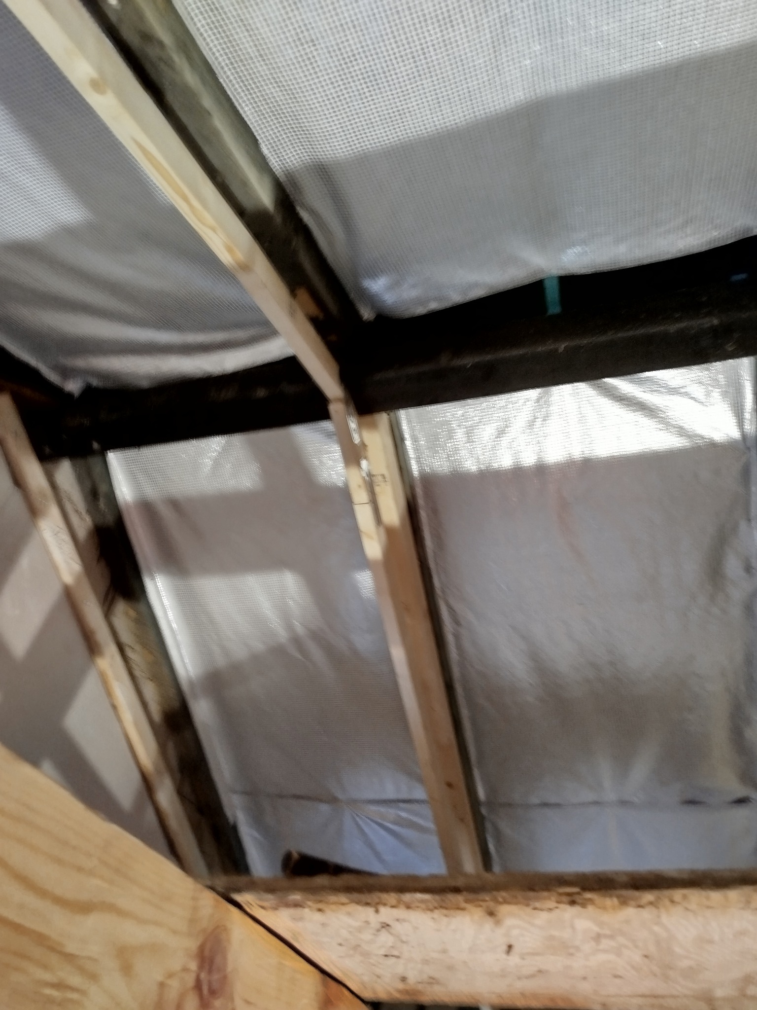 Skośny dach z drewnianą konstrukcją i folią paroizolacyjną, widok z dołu. Elementy drewniane surowe, nieobrobione. Detal montażu izolacji.