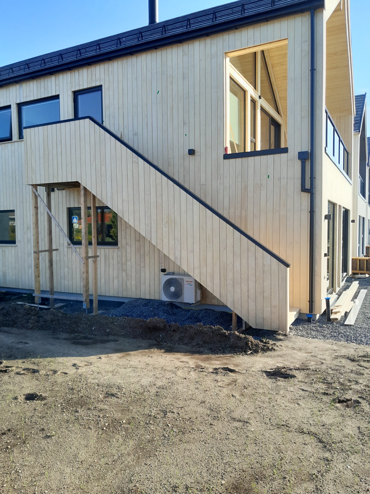 Jasny dom szkieletowy z drewnianą elewacją i zewnętrznymi schodami. Widoczny klimatyzator i tymczasowe podparcie nad wejściem. Błękitne niebo w tle.