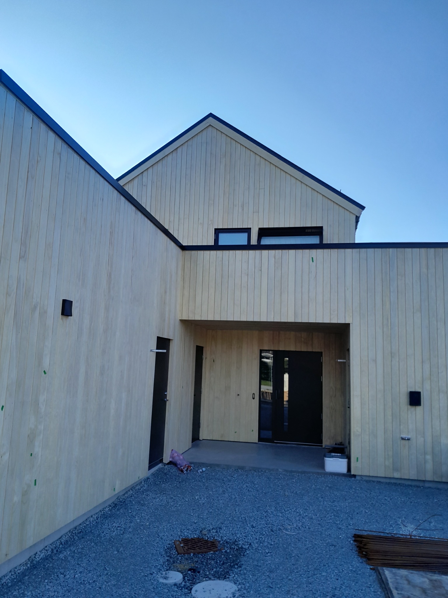 Nowoczesny dom szkieletowy z jasną elewacją z desek, czarnymi ramami okien i drzwiami. Na pierwszym planie żwirowe podłoże i elementy wykończeniowe.