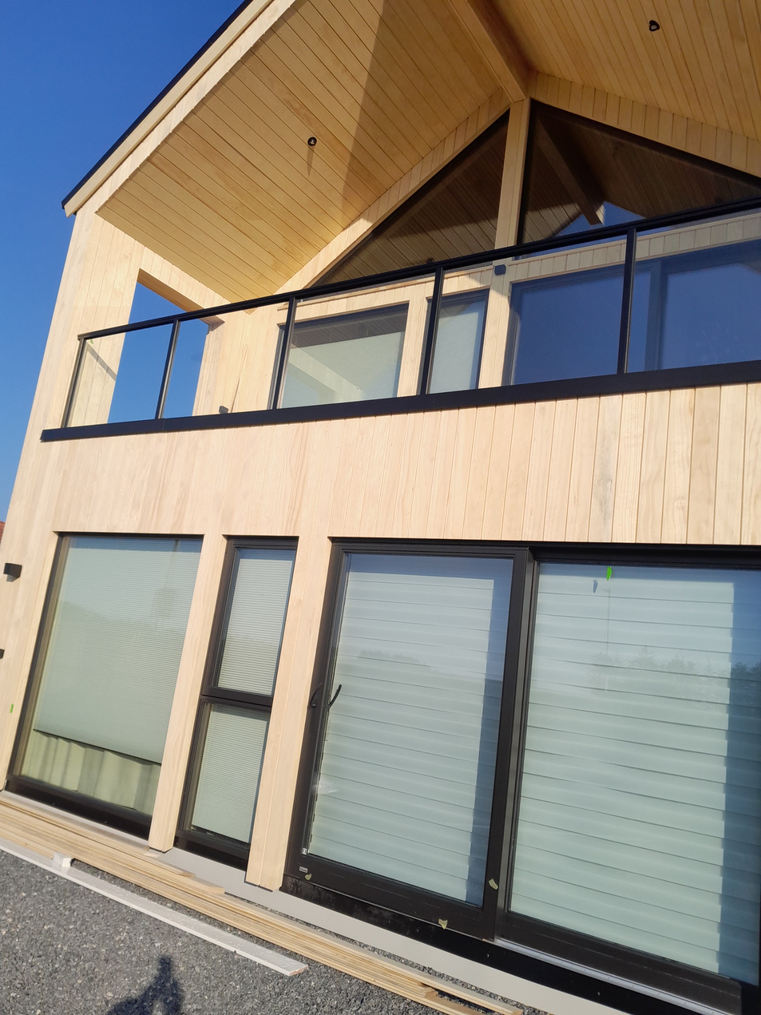 Nowoczesny dom szkieletowy z drewnianą elewacją i dużymi oknami, balkon z czarną balustradą. Jasne drewno i minimalistyczny design.