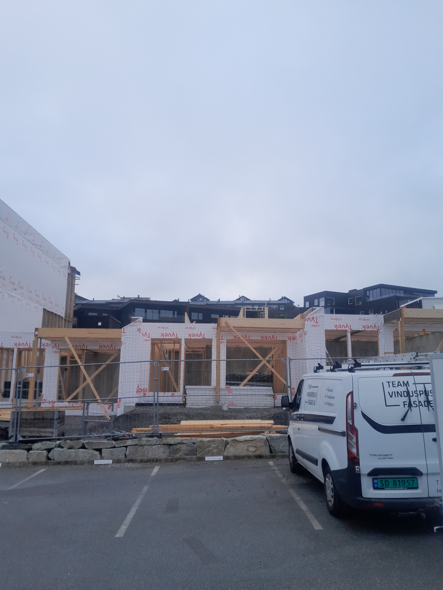 Wznoszenie domu szkieletowego w trakcie budowy, widoczne drewniane ramy i biała membrana. Na pierwszym planie zaparkowany biały bus firmy budowlanej.