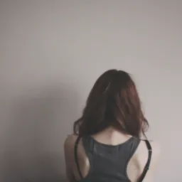 Kobieta z długimi, rudymi włosami, ubrana w szary top na ramiączkach, stoi tyłem na tle jasnej ściany. Delikatne światło podkreśla naturalny kolor włosów.