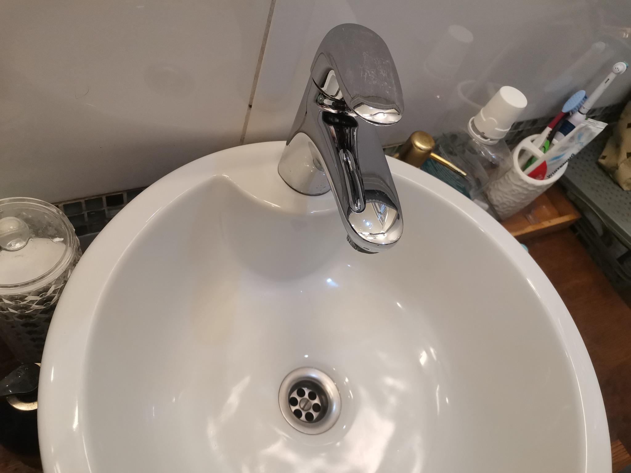 Biała umywalka z chromowaną baterią, widoczne akcesoria łazienkowe i osady kamienia na armaturze.