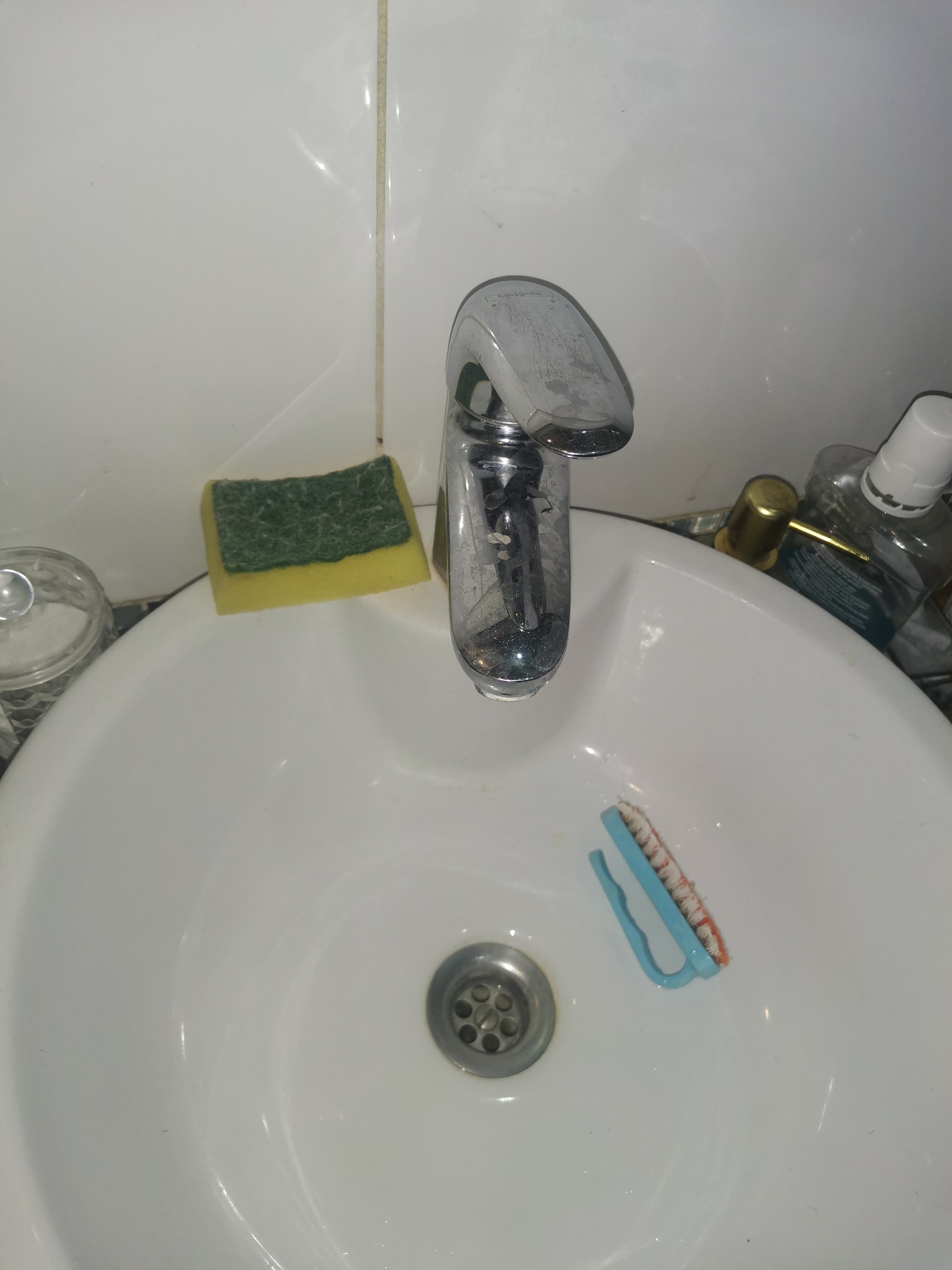 Umywalka z chromowaną baterią, gąbką, szczotką do czyszczenia i butelkami z płynami, wymagająca sprzątania.