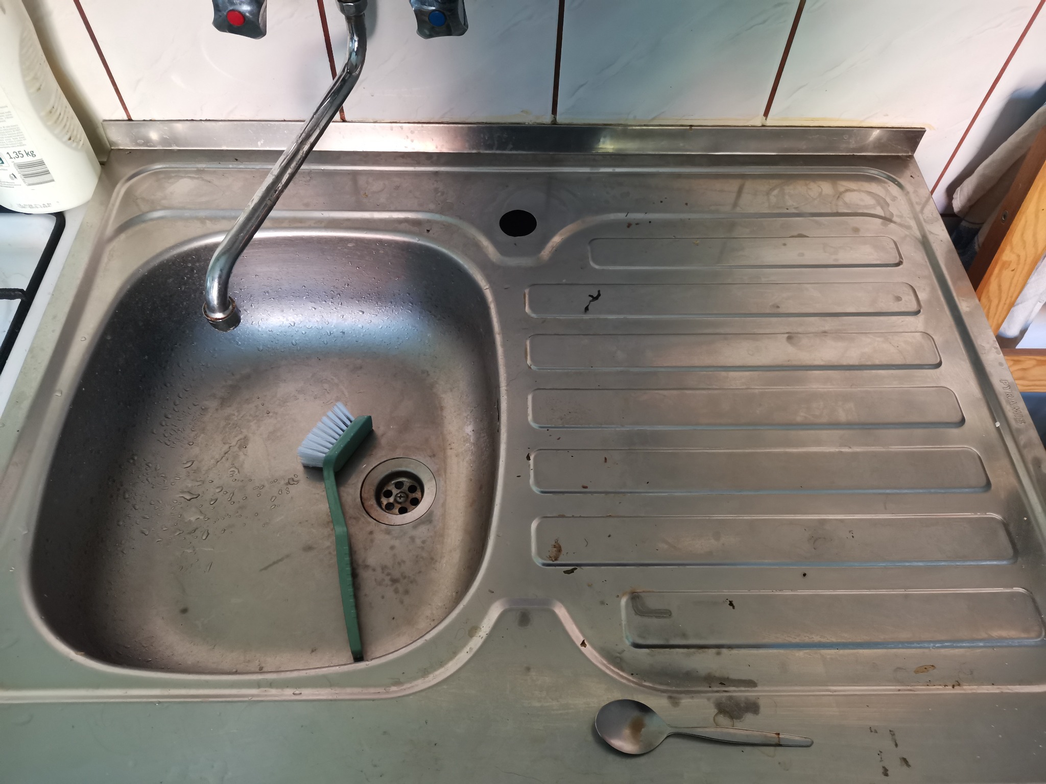 Używany, stalowy zlew kuchenny z widocznymi śladami użytkowania, szczotką do szorowania i łyżeczką. Widoczny kran i fragment płytek ściennych.
