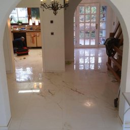 HOME INVESTING Aleksander Zera - Błyszcząca, kremowa podłoga z płytek imitujących marmur, ułożona w przedpokoju z łukowatymi przejściami; w tle widoczna osoba pracująca w kuchni i wiszący żyrandol.