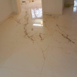 HOME INVESTING Aleksander Zera - Układanie beżowych płytek podłogowych z marmurkowym wzorem, widoczny majster z narzędziami w tle.