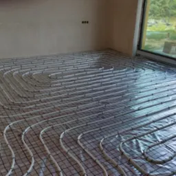 Rozłożone rury ogrzewania podłogowego na siatce w surowym wnętrzu, widoczne przyłącza na ścianie i okno w tle.