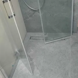 Szara łazienka z kabiną prysznicową typu walk-in, widoczny odpływ liniowy i płytki imitujące kamień. Minimalistyczny design, nowoczesne wykończenie.