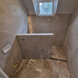 HOME INVESTING Aleksander Zera - Wykończona łazienka w beżowych płytkach imitujących marmur, z oknem w skosie. Widoczne przygotowane instalacje wodne i elektryczne, oraz fragment niedokończonej podłogi.