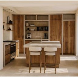 HOME INVESTING Aleksander Zera - Jasna kuchnia z drewnianymi szafkami, wyspą z kamiennym blatem i dwoma krzesłami barowymi. Minimalistyczny design, ciepłe oświetlenie, płytki na ścianie.