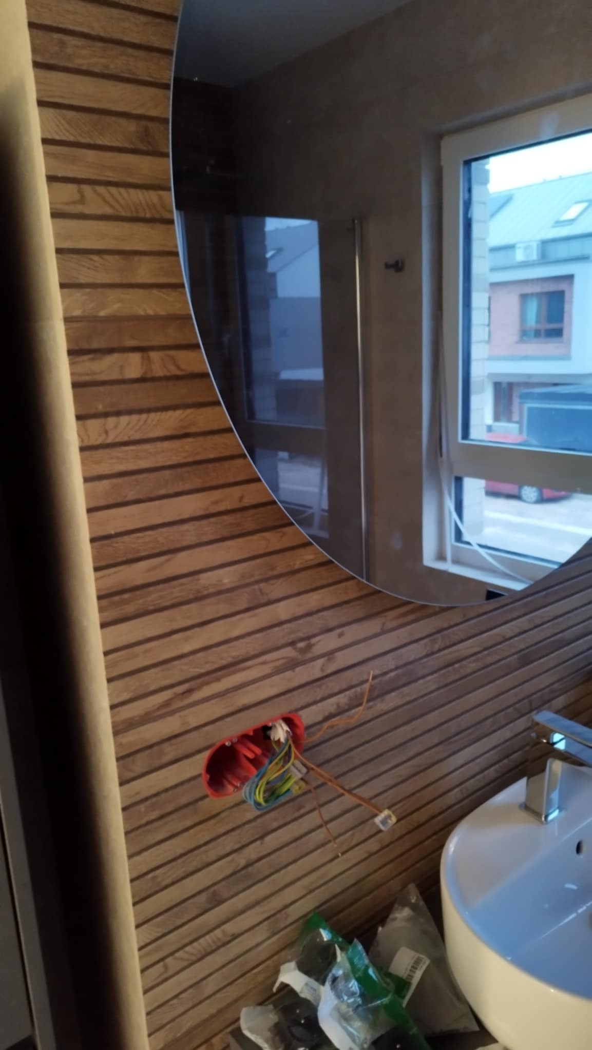 Niedokończona instalacja elektryczna w łazience z drewnopodobną okładziną ściany, widoczna puszka elektryczna z kablami, umywalka i lustro z odbiciem okna.