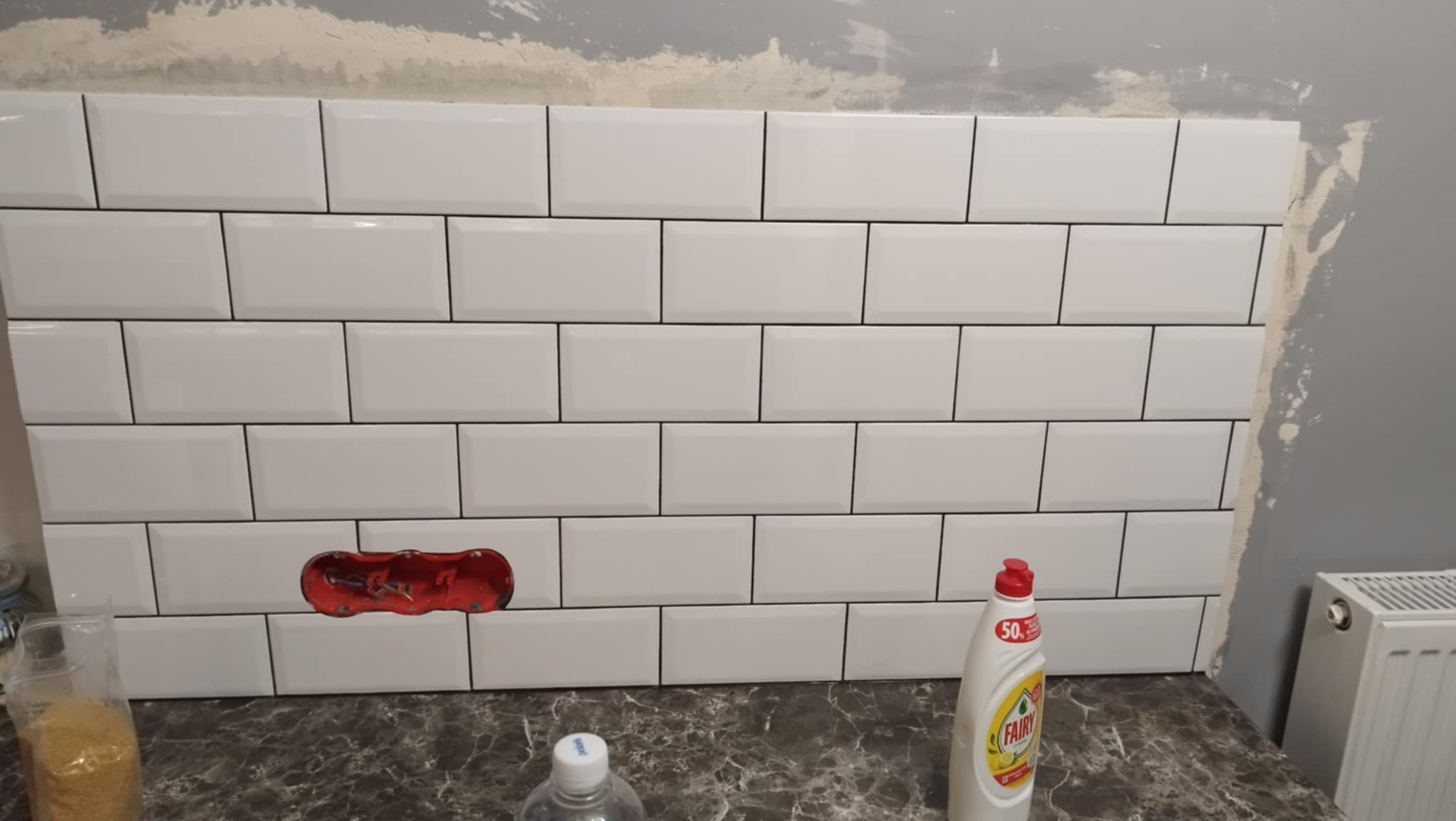 Ściana z białych, prostokątnych płytek ułożonych w cegiełkę, z widocznym gniazdkiem elektrycznym w trakcie montażu i butelką płynu do naczyń Fairy na blacie.