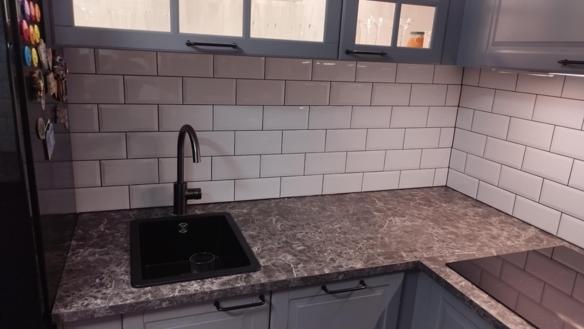Szare szafki kuchenne z marmurkowym blatem i białymi płytkami ułożonymi w cegiełkę, czarny zlew i bateria w Ostrowi Mazowieckiej.