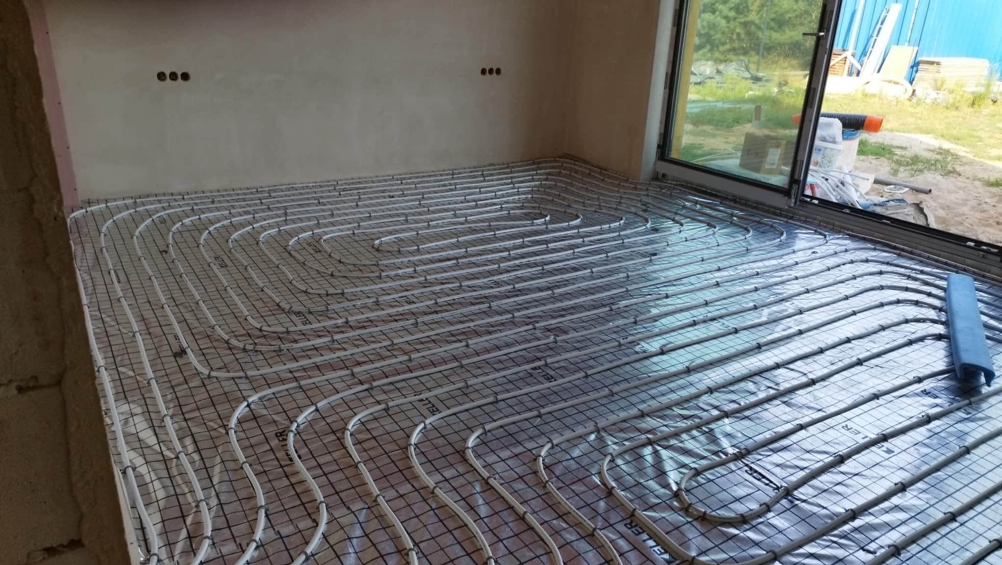 Rozłożone rury ogrzewania podłogowego na siatce w surowym wnętrzu, widoczne przyłącza na ścianie i okno w tle.