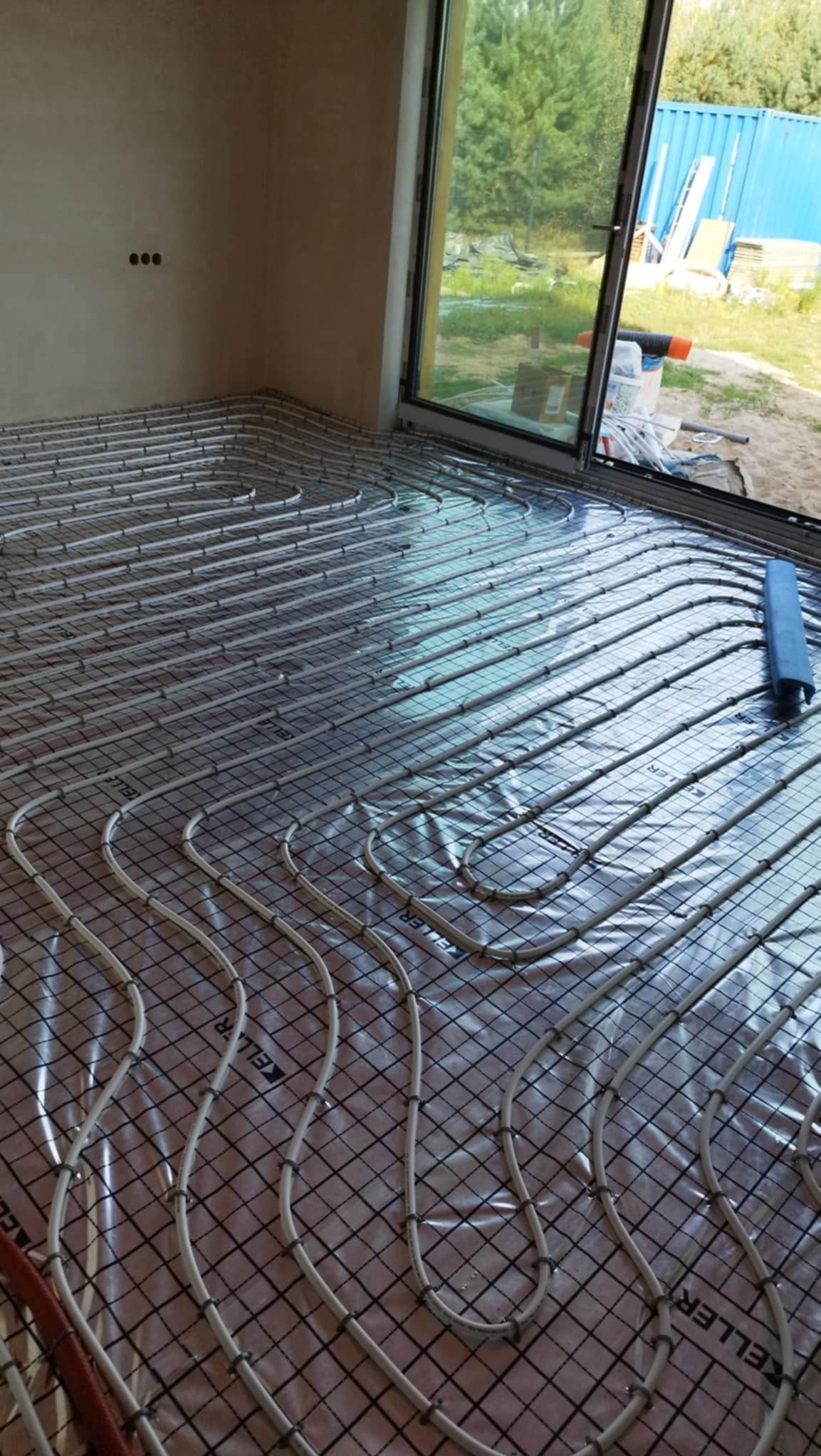 Instalacja ogrzewania podłogowego z rur na folii odblaskowej, widoczne mocowania i narzędzie do montażu, surowy stan ścian, w tle okno z widokiem na zewnątrz.