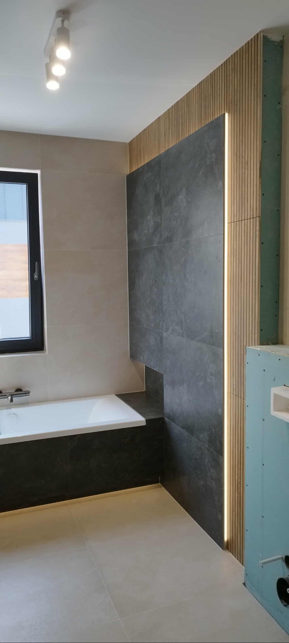 Nowoczesna łazienka po remoncie: szare płytki, drewniane lamele z oświetleniem LED, wanna z obudową w kolorze grafitu, jasna podłoga i czarne okno.