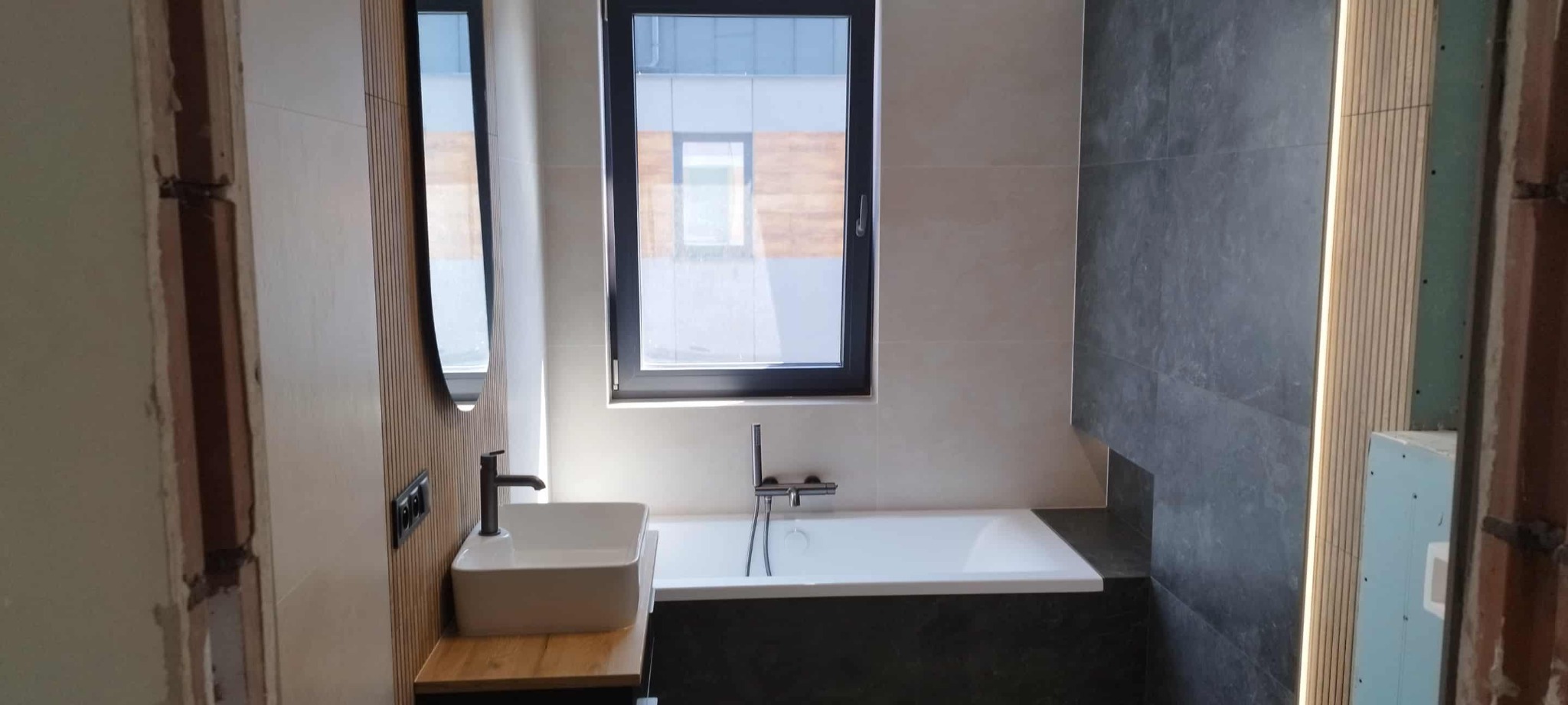 Nowoczesna łazienka w trakcie wykańczania: biała wanna, umywalka na drewnianym blacie, szare płytki, drewniane panele ścienne i czarne okno. Widoczne niedokończone fragmenty.