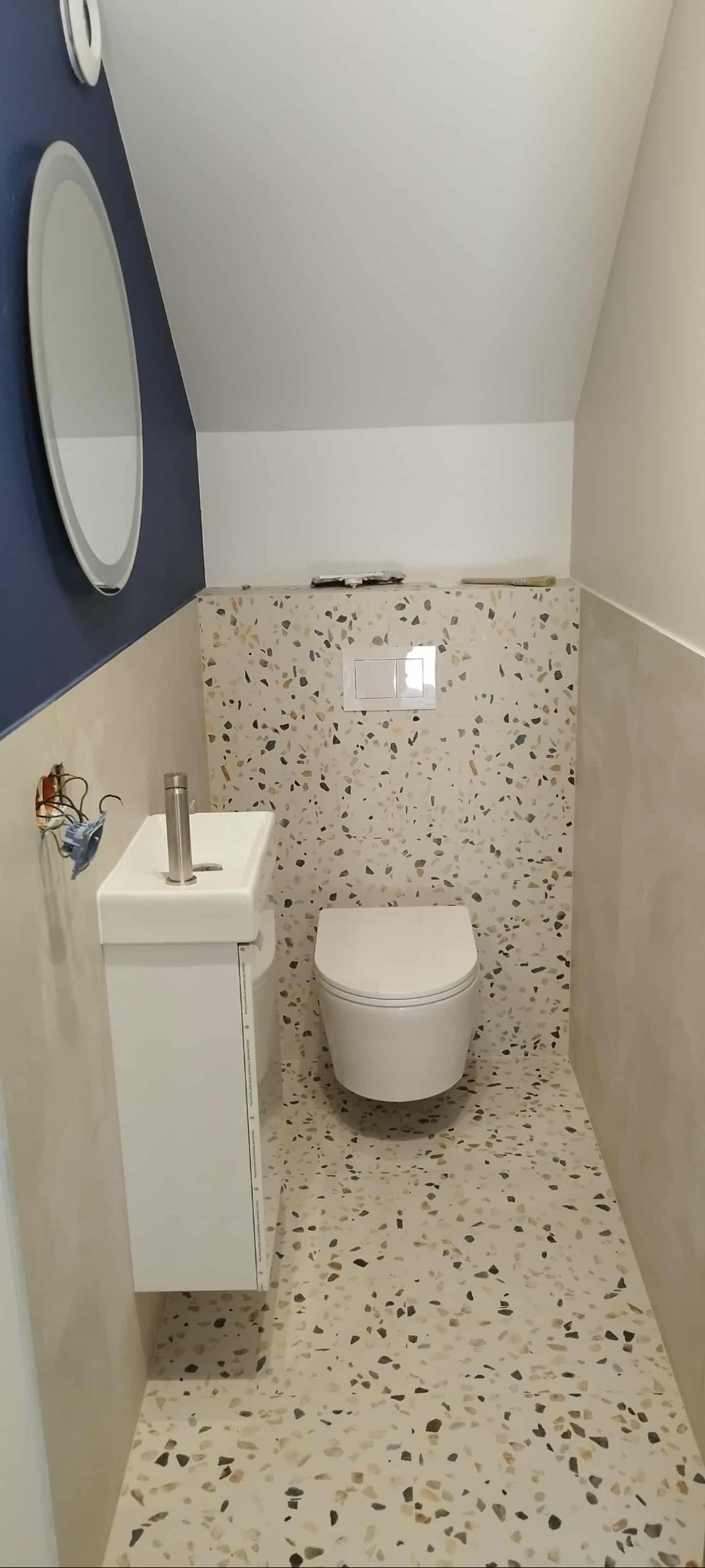 Nowoczesna łazienka z umywalką na szafce, wiszącą toaletą i mozaikową okładziną ścian i podłogi. Niebieski akcent kolorystyczny na ścianie z lustrem.