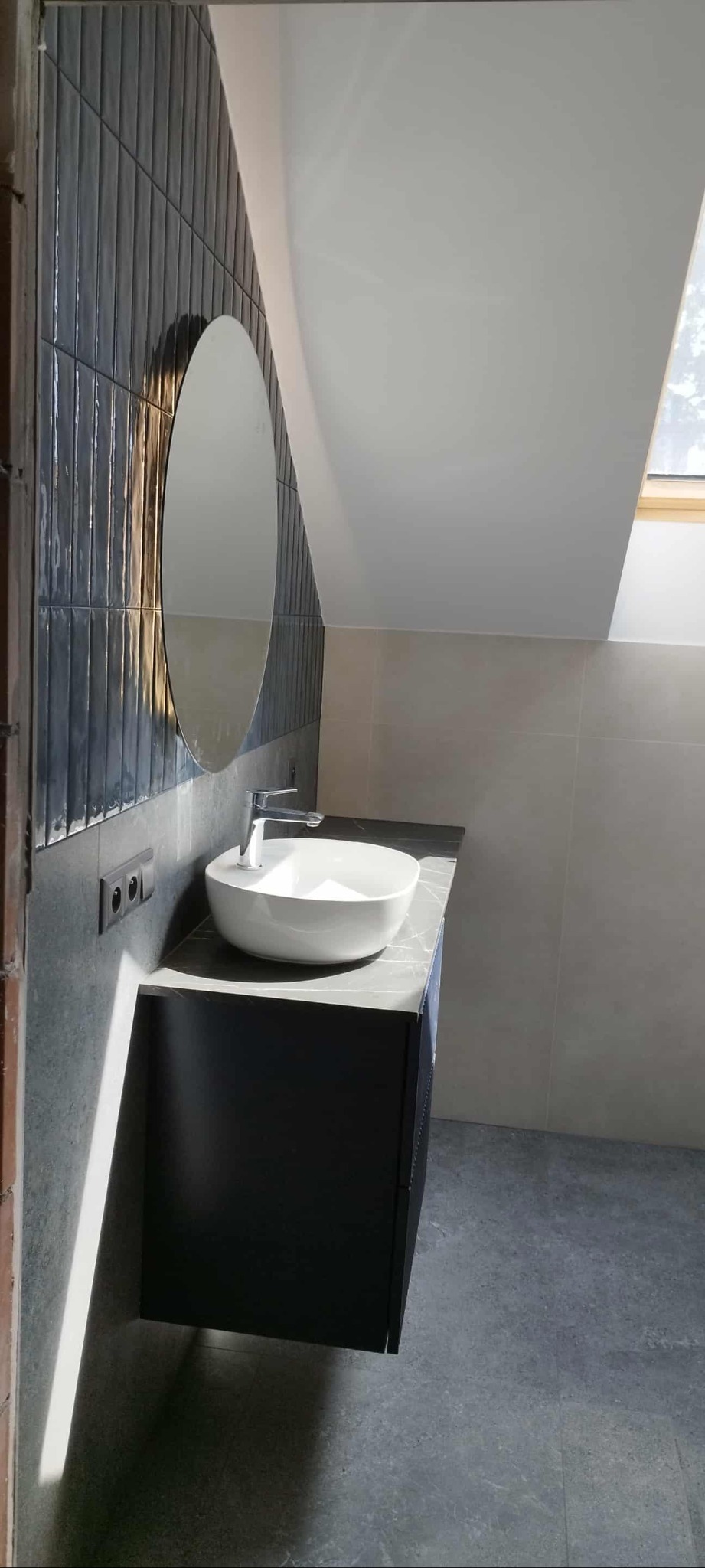 Nowoczesna łazienka na poddaszu z owalnym lustrem, umywalką nablatową i grafitowymi płytkami imitującymi cegłę. Światło słoneczne wpada przez okno dachowe.