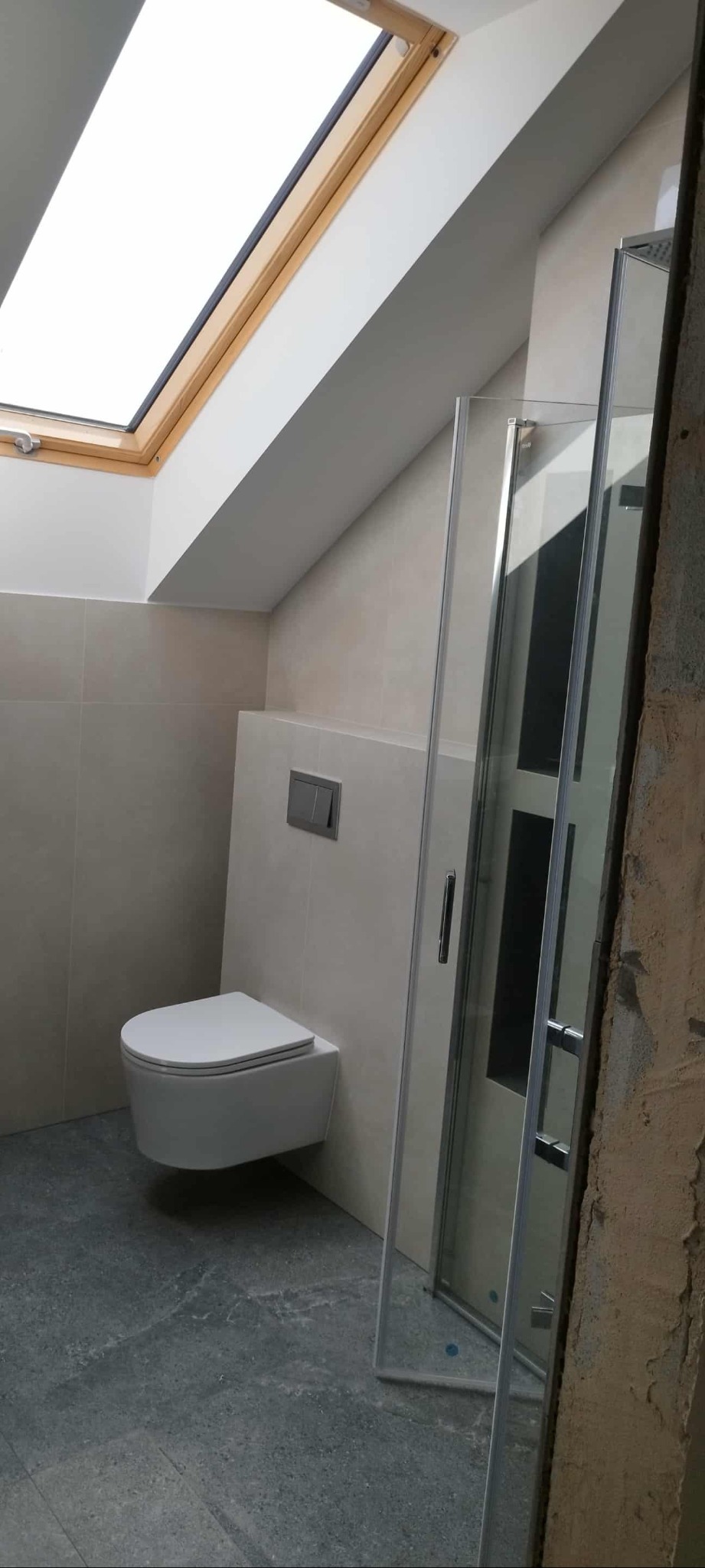 Nowoczesna łazienka na poddaszu z oknem dachowym. Wisząca toaleta, szklana kabina prysznicowa i beżowe płytki tworzą minimalistyczny design.