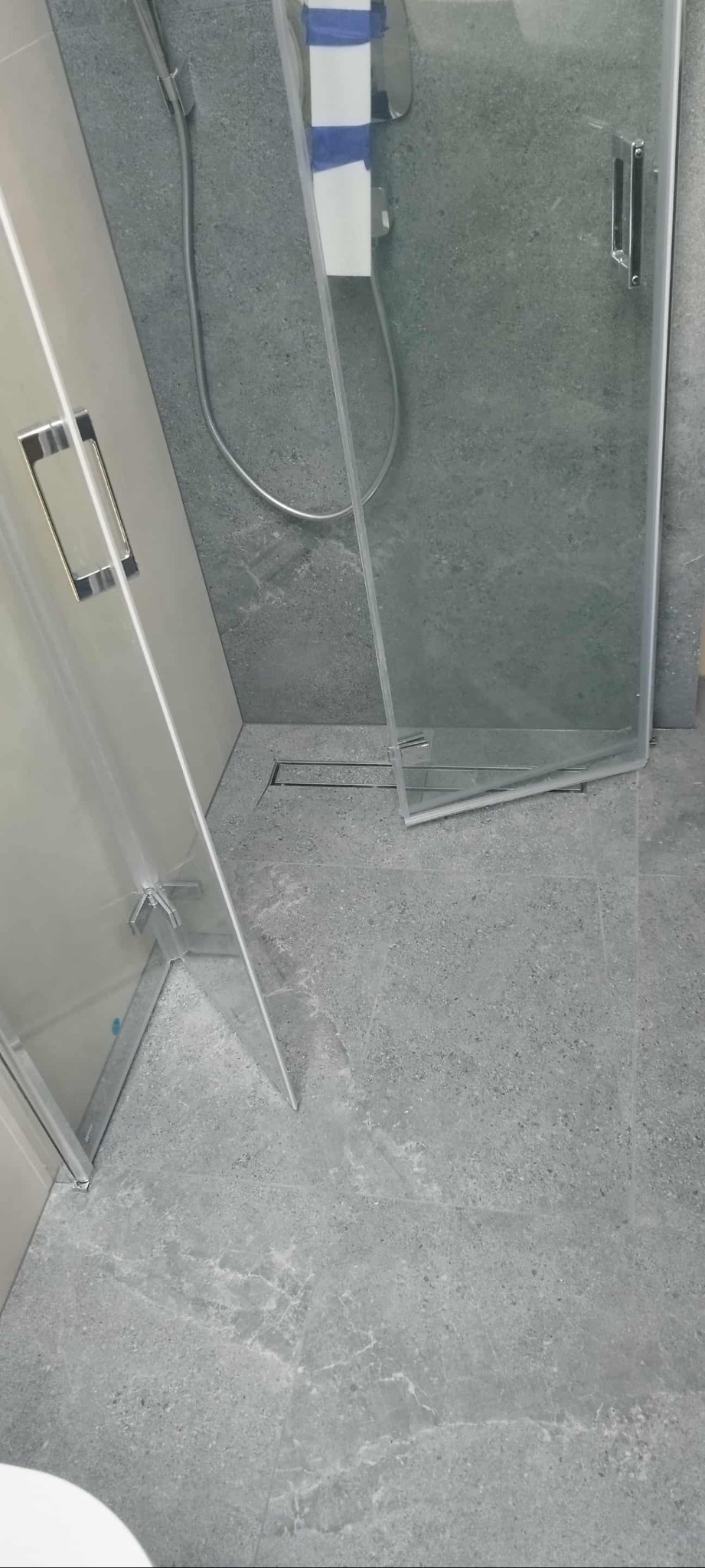 Szara łazienka z kabiną prysznicową typu walk-in, widoczny odpływ liniowy i płytki imitujące kamień. Minimalistyczny design, nowoczesne wykończenie.