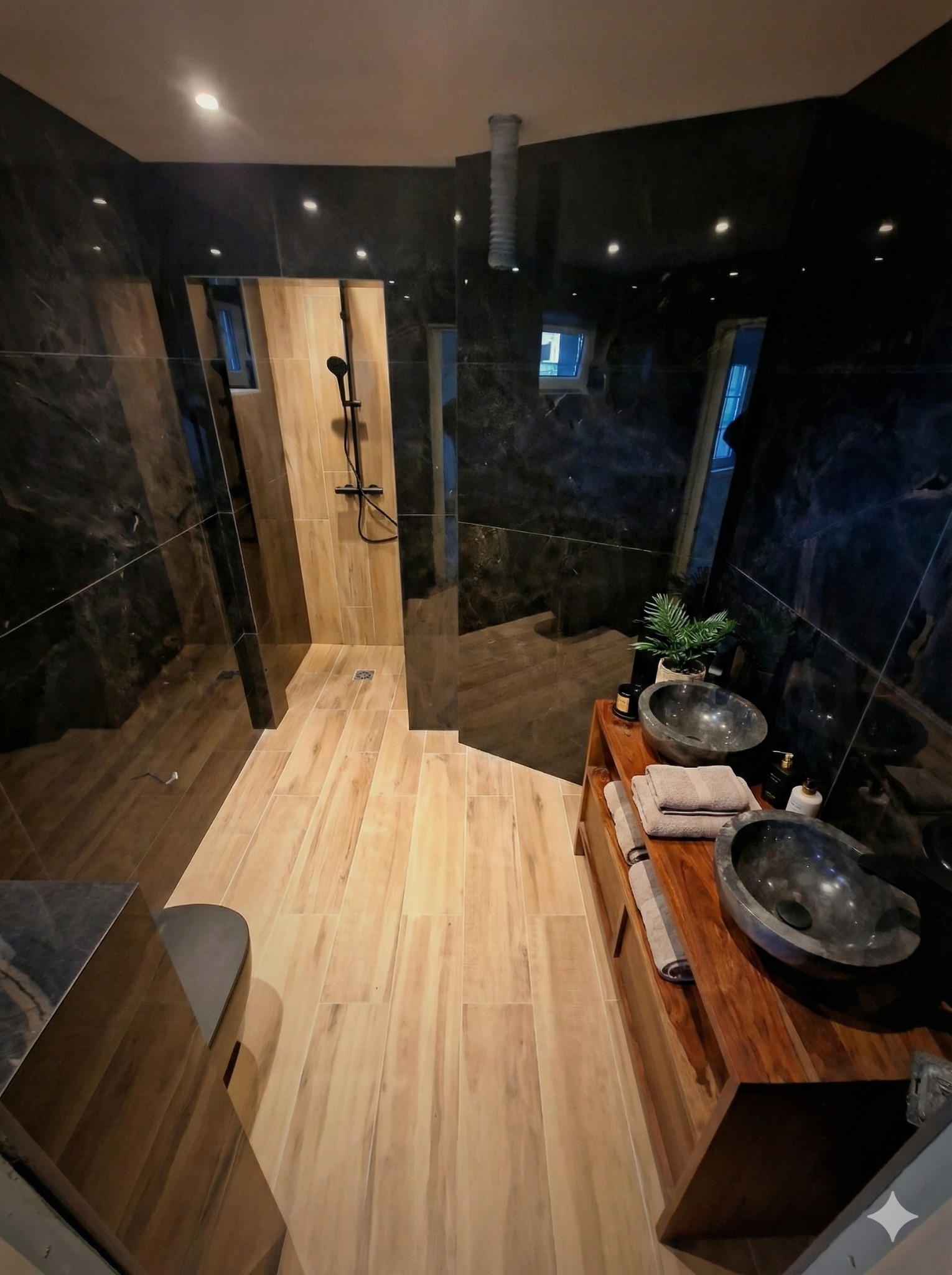 Nowoczesna łazienka z kabiną prysznicową typu walk-in, drewnianą podłogą i umywalkami z kamienia na drewnianej szafce. Czarne marmurowe ściany i oświetlenie punktowe.